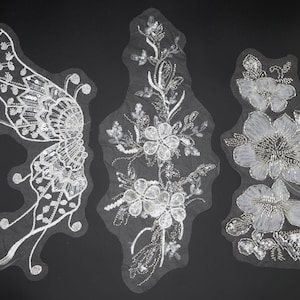 Peut inclure: Trois appliques brodées blanches distinctes sur un support en maille transparente. La pièce de gauche a une forme d'aile de papillon, celle du centre est une vigne florale verticale et celle de droite présente trois grandes fleurs, ornées de sequins argentés.