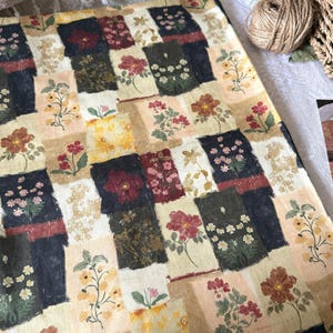 May include: A fabric featuring a patchwork design of rectangular blocks in shades of cream, beige, and dark navy. Each block displays various floral illustrations in red, yellow, white, and green, creating a repeating botanical pattern across the textile.