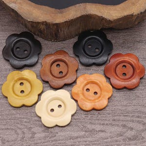 May include: A selection of flower-shaped wooden buttons in shades of black, brown, and cream. Each button features two sewing holes. The buttons are displayed on a wooden surface.