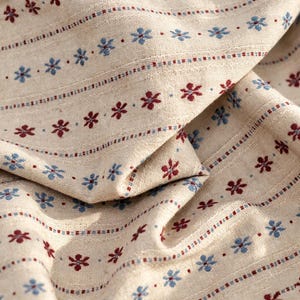 Könnte beinhalten: Ein beigefarbener Webstoff mit einem sich wiederholenden Muster aus kleinen blauen und dunkelroten Blumen in horizontalen Reihen. Das Textil hat eine sichtbare Webstruktur mit dünnen vertikalen Streifen aus blauen und roten Punkten.
