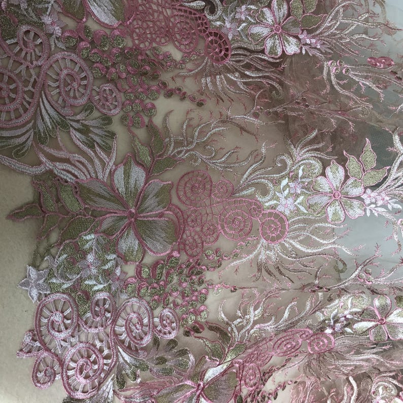 May include: Close-up of a delicate floral lace fabric. The design features embroidered flowers, leaves, and swirling patterns in shades of pink, white, and gold. The lace has a sheer, airy quality, perfect for apparel or craft projects.