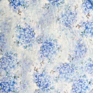 May include: A fabric featuring a repeating pattern of blue hydrangea flower clusters on a white background. The design includes soft watercolour-style petals in various shades of blue, interspersed with small green leaves and thin brown stems.