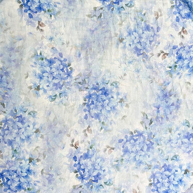 May include: A fabric featuring a repeating pattern of blue hydrangea flower clusters on a white background. The design includes soft watercolour-style petals in various shades of blue, interspersed with small green leaves and thin brown stems.