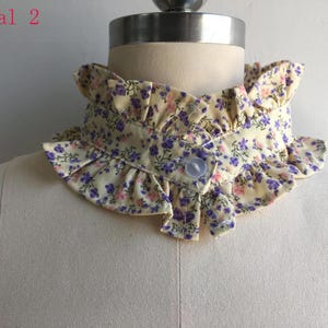 May include: A detachable collar featuring a pale yellow fabric with a small purple and pink floral print. The collar has ruffled edges along the top and bottom, a front button closure, and the text floral 2 printed in the upper left corner.
