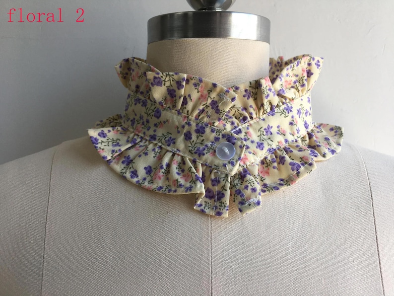 May include: A detachable collar featuring a pale yellow fabric with a small purple and pink floral print. The collar has ruffled edges along the top and bottom, a front button closure, and the text floral 2 printed in the upper left corner.