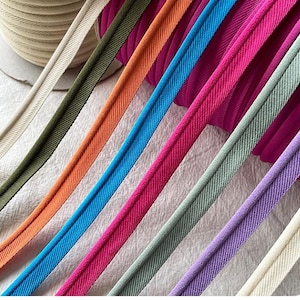 Puede incluir: Primer plano de cinta de bies colorida en varios tonos, incluyendo verde, naranja, azul, rosa y morado. La cinta está cuidadosamente dispuesta, mostrando su textura y colores vibrantes. Un rollo de cinta beige es visible en el fondo, sugiriendo un tema de costura o artesanía.