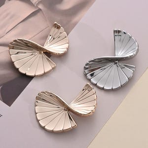 May include: Three abstract, fan-shaped earrings in gold and silver tones. The earrings have a spiral design with multiple, thin, curved lines. The earrings are displayed on a neutral background.