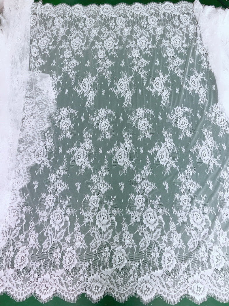 May include: White floral lace fabric with a delicate pattern. The lace has a scalloped edge and is sheer and lightweight.