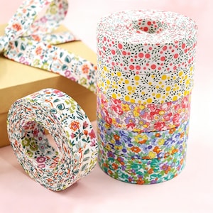 Könnte beinhalten: Ein Stapel aus fünf Stoffbandrollen mit verschiedenen Blumenmustern in Rosa, Gelb, Rot, Blau und Grün, neben einer einzelnen passenden Rolle und einer beigen Schachtel, die mit demselben floral gemusterten Stoffband umwickelt ist.