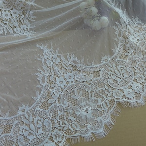 Peut inclure: Gros plan sur un tissu de dentelle blanche délicate avec des motifs floraux et de feuilles. La dentelle a un bord festonné et un fond à pois, créant une texture douce et élégante. Idéal pour les tenues de mariée ou les projets d'artisanat.