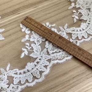 May include: Close-up of white floral lace trim with a wooden ruler measuring up to 21 cm. The lace features detailed flower and leaf patterns. The background is a light brown wooden surface. Ideal for sewing and craft projects.