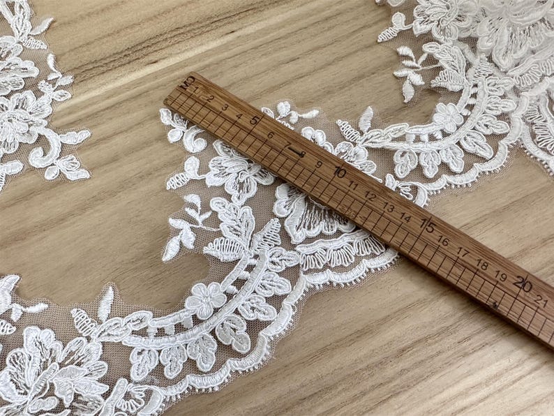 May include: Close-up of white floral lace trim with a wooden ruler measuring up to 21 cm. The lace features detailed flower and leaf patterns. The background is a light brown wooden surface. Ideal for sewing and craft projects.