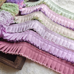 Puede incluir: Surtido de cintas de tela plisadas en varios colores pastel, incluyendo rosa, lavanda y verde. Cada cinta presenta un borde festoneado y costuras decorativas. Las cintas están dispuestas en una bandeja de madera, mostrando sus texturas y tonos.