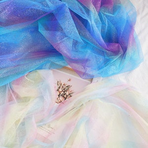 Könnte beinhalten: Transparenter Tüllstoff mit einem Farbverlauf in Blau- und Violetttönen mit silbernen Glitzerakzenten, der über einem zweiten Stück irisierendem Pastell-Tüll liegt, welches eine kleine rechteckige Karte mit Blumenmotiv teilweise verdeckt.
