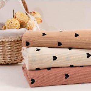 Puede incluir: Una pila de tres telas de pana en tonos beige, crema y rosa, cada una adornada con patrones de corazones negros. Las telas están cuidadosamente dobladas, mostrando su textura y diseño. Una cesta de mimbre con flores amarillas en el fondo.
