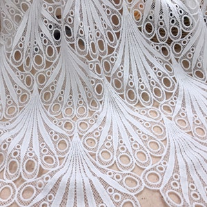 May include: White lace fabric with a detailed peacock feather design. The fabric features a repeating pattern of open circles and curved lines, creating a delicate and airy texture. Ideal for dresses, skirts, or decorative projects.