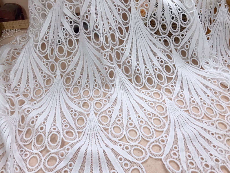 May include: White lace fabric with a detailed peacock feather design. The fabric features a repeating pattern of open circles and curved lines, creating a delicate and airy texture. Ideal for dresses, skirts, or decorative projects.