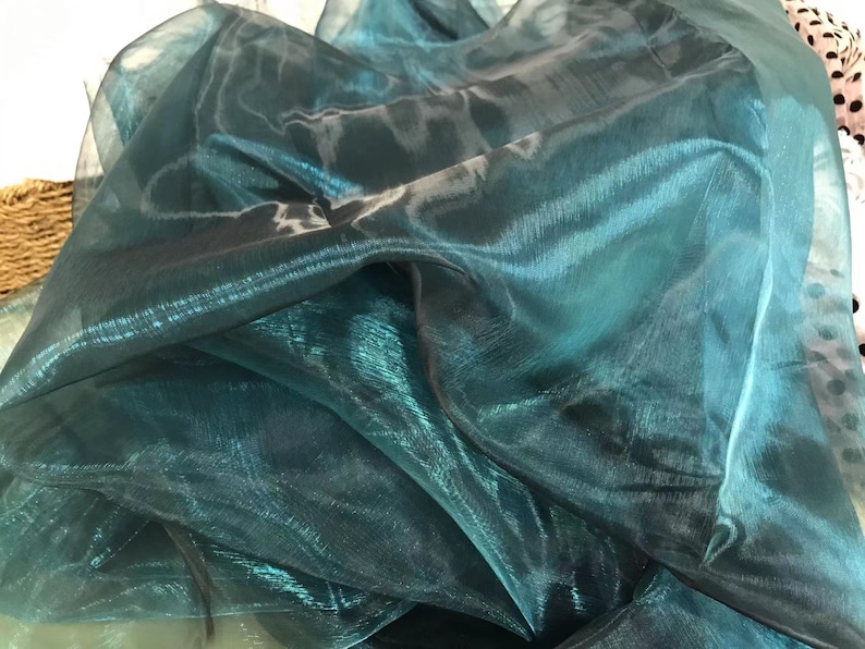 May include: Teal iridescent sheer fabric with a shimmery finish. The fabric is folded and draped, showing its texture and drape.