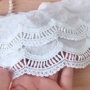 May include: White cotton lace trim with a scalloped edge and a delicate crochet design. The lace has two layers, with the top layer having a more open design than the bottom layer.