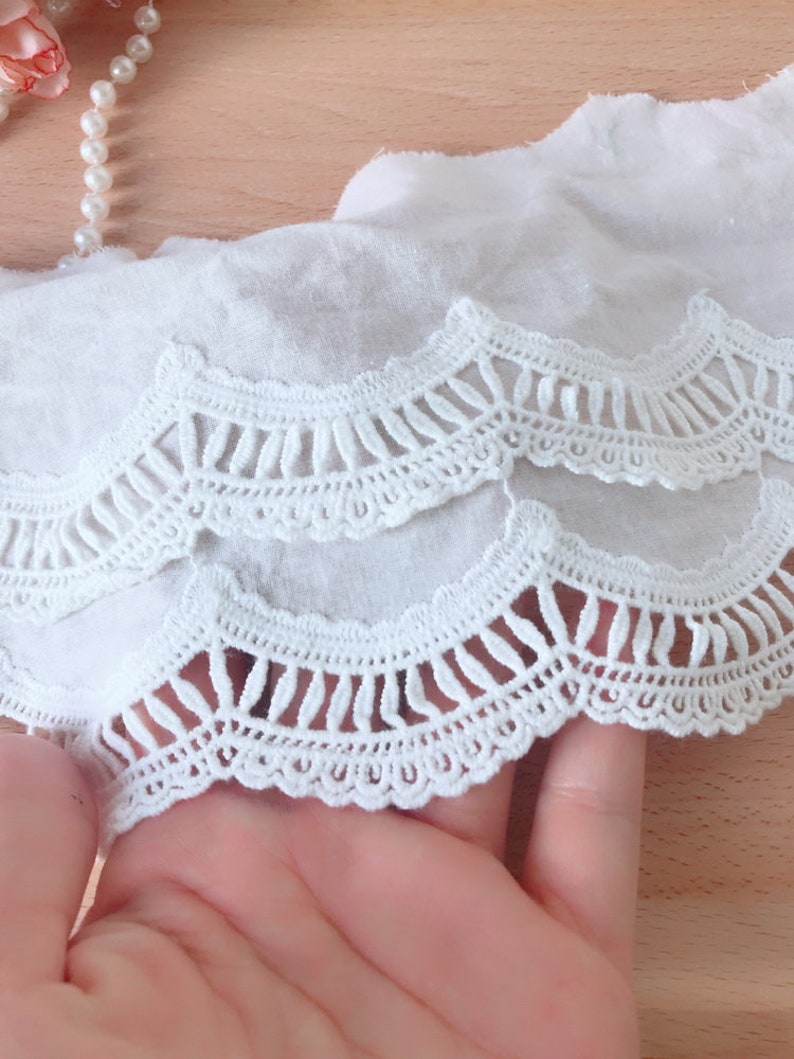 May include: White cotton lace trim with a scalloped edge and a delicate crochet design. The lace has two layers, with the top layer having a more open design than the bottom layer.