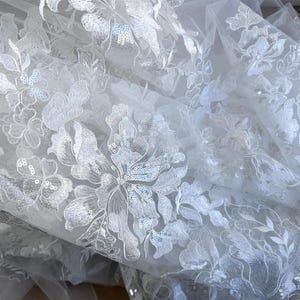 May include: White floral lace fabric with sequins. The lace has a delicate floral pattern and is perfect for adding a touch of elegance to any project.