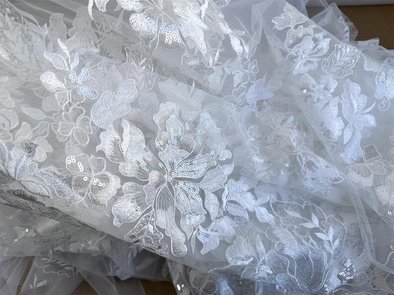 May include: White floral lace fabric with sequins. The lace has a delicate floral pattern and is perfect for adding a touch of elegance to any project.
