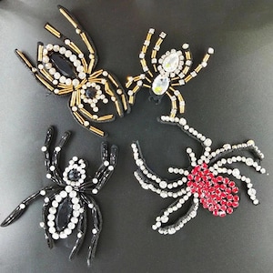 Peut inclure: Quatre écussons décoratifs en forme d'araignée ornés de perles et de strass. Une araignée présente des perles dorées et des pierres transparentes, une autre des pierres transparentes, une des perles noires et une des pierres rouges et claires.