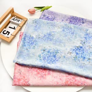May include: Three folded pieces of fabric featuring floral patterns in shades of purple, light blue, and pink. The fabric has a textured appearance with hydrangea-like flower prints. A wooden calendar block displays the text APR 15.