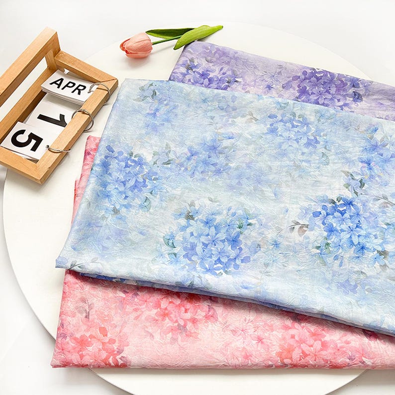 May include: Three folded pieces of fabric featuring floral patterns in shades of purple, light blue, and pink. The fabric has a textured appearance with hydrangea-like flower prints. A wooden calendar block displays the text APR 15.