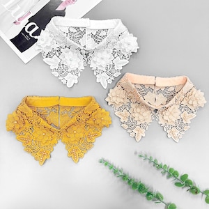 May include: Three lace collar accessories in white, mustard yellow, and beige. Each collar features floral and leaf designs with pearl accents. The collars are displayed on a light grey surface.