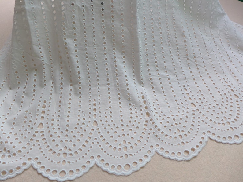 May include: White cotton fabric with a scalloped edge and a repeating pattern of small circles cut out. The fabric is delicate and airy.