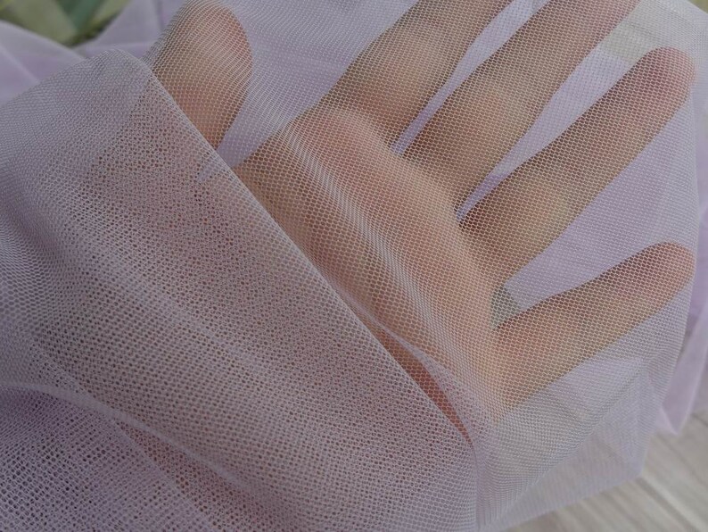 May include: A close-up of a pale lavender sheer fabric with a fine mesh texture. The fabric is soft and delicate, with a slightly iridescent sheen.