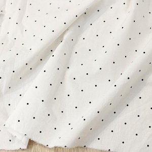 May include: A white fabric featuring a repeating pattern of small, evenly spaced black polka dots. The material is draped in soft folds, highlighting the texture of the textile and the consistent distribution of the dark circular print across the surface.