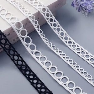 Può includere: Primo piano di diversi nastri decorativi. Un nastro nero e due nastri bianchi con un motivo a cerchi ripetuti sono visibili. I nastri sono realizzati in un materiale morbido e strutturato. Adatto per artigianato o abbellimenti.