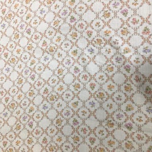 May include: Cream-colored fabric with a repeating pattern of small, colorful flowers. The flowers are arranged in a grid-like pattern, with each flower cluster surrounded by a delicate, linked design. The flowers are in shades of pink, yellow, and purple.