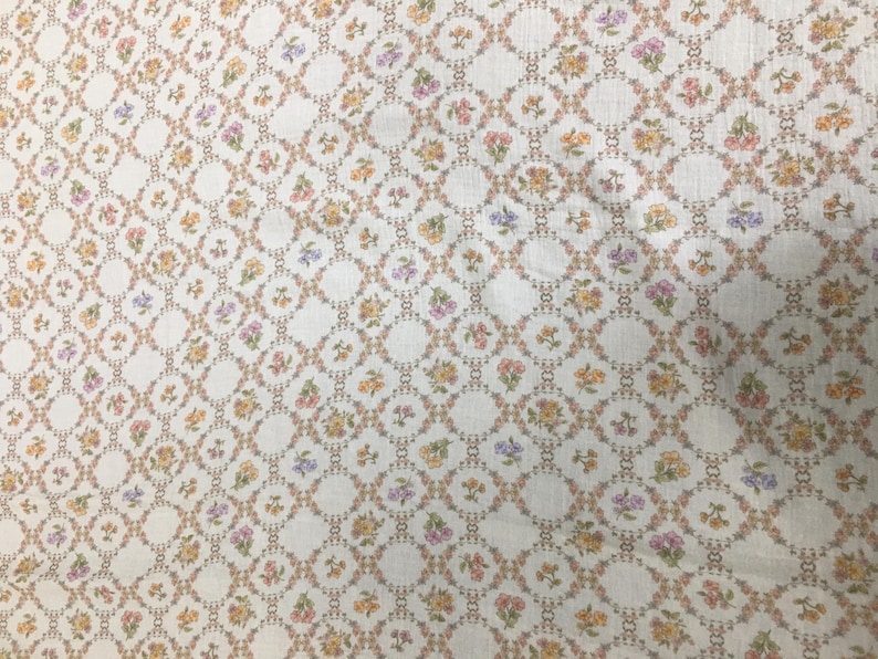 May include: Cream-colored fabric with a repeating pattern of small, colorful flowers. The flowers are arranged in a grid-like pattern, with each flower cluster surrounded by a delicate, linked design. The flowers are in shades of pink, yellow, and purple.