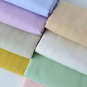 May include: A stack of seven fabric swatches in various pastel colors, each with a delicate, repeating lace pattern. The fabrics are all ribbed and have a soft, textured feel.