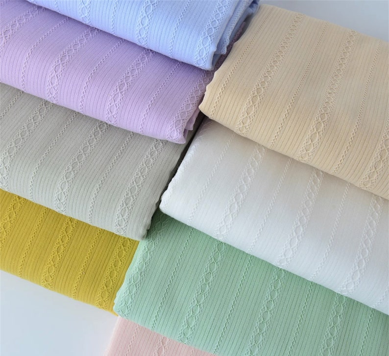 May include: A stack of seven fabric swatches in various pastel colors, each with a delicate, repeating lace pattern. The fabrics are all ribbed and have a soft, textured feel.