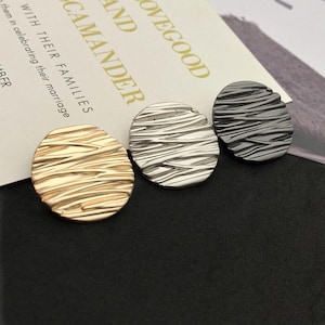 May include: Three textured, oval-shaped metal buttons in gold, silver, and dark gray. The buttons have a ridged pattern and are displayed on a black surface with a wedding invitation in the background.