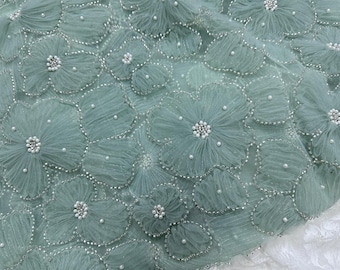 May include: A close-up of a light blue fabric with 3D floral embellishments. The fabric is covered in small, white pearls and has a delicate, airy texture.