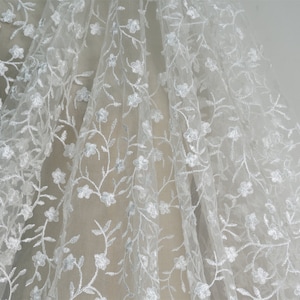 May include: White floral lace fabric featuring a delicate pattern of small flowers and vines. The lace is sheer and has a soft, flowing texture.