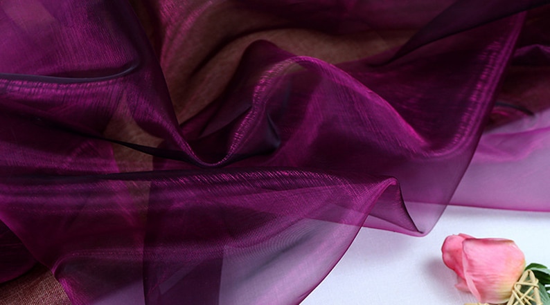 May include: A close-up of a sheer, purple fabric with a slight sheen. The fabric is draped and folded, creating soft, flowing lines.
