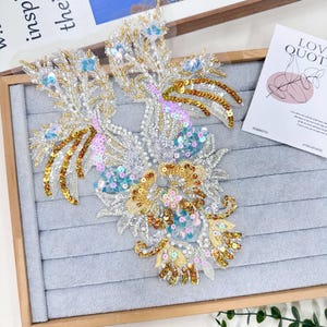 Może przedstawiać: Ozdobna, haftowana aplikacja z kwiatowym wzorem. Aplikacja posiada złote, srebrne i niebieskie cekiny i koraliki. Projekt znajduje się na szarym tle, z drewnianą ramą. Karta z napisem "LOVE QUOTE" znajduje się w tle.