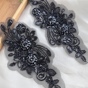 Könnte beinhalten: Zwei schwarze Blumen-Applikationen. Jede Applikation weist aufwendige Perlenarbeiten, Pailletten und gestickte Details auf. Das Design umfasst Blumen, Blätter und wirbelnde Muster, alles auf einem transparenten schwarzen Netzrücken. Ideal zum Verzieren von Kleidungsstücken oder Accessoires.