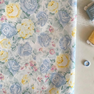 May include: A close-up of a floral fabric featuring a pattern of blue, yellow, and pink roses on a white background. The roses are surrounded by green leaves. The fabric is folded, with spools of thread and a button in the background.