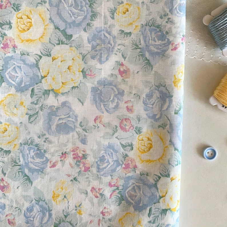 May include: A close-up of a floral fabric featuring a pattern of blue, yellow, and pink roses on a white background. The roses are surrounded by green leaves. The fabric is folded, with spools of thread and a button in the background.