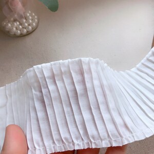 K&ouml;nnte beinhalten: Wei&szlig;er gefalteter Stoffbesatz mit einem gen&auml;hten Rand. Der Besatz ist in der Mitte gefaltet und zeigt die Falten auf beiden Seiten.