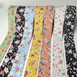 Könnte beinhalten: Eine Sammlung von Bändern mit floralen Mustern in verschiedenen Farben, darunter Grün, Schwarz, Hellblau, Gelb, Orange, Weiß und Schwarz. Jedes Band hat ein anderes Blumendesign mit weißen Kanten. Die Bänder sind nebeneinander angeordnet.
