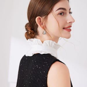 May include: A sleeveless black dress with a white ruffled collar. The dress is embellished with small, shiny sequins. The model is wearing silver earrings. The background is plain white.