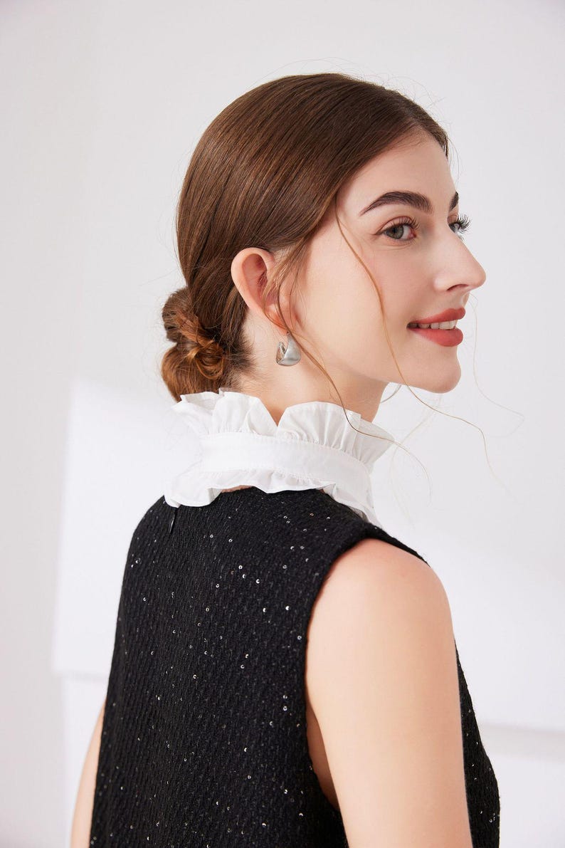 May include: A sleeveless black dress with a white ruffled collar. The dress is embellished with small, shiny sequins. The model is wearing silver earrings. The background is plain white.